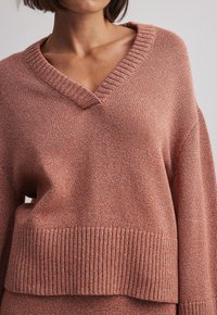 Pull en maille couleur rouille avec col en V, poignets et ourlet côtelés. Texture douce et coupe décontractée, avec un motif tissé sur toute la surface.