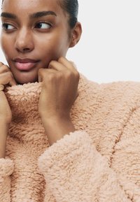 Pink fleecejakke med høj krave, blød tekstur og opsmøgede ærmekanter, hænder holder kraven nær ansigtet.