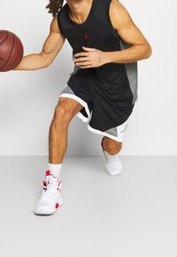 Camiseta de tirantes negra con logo rojo, detalles laterales grises y pantalones cortos negros a juego. Lleva zapatillas de baloncesto blancas y rojas. Sostiene una pelota en la mano derecha.