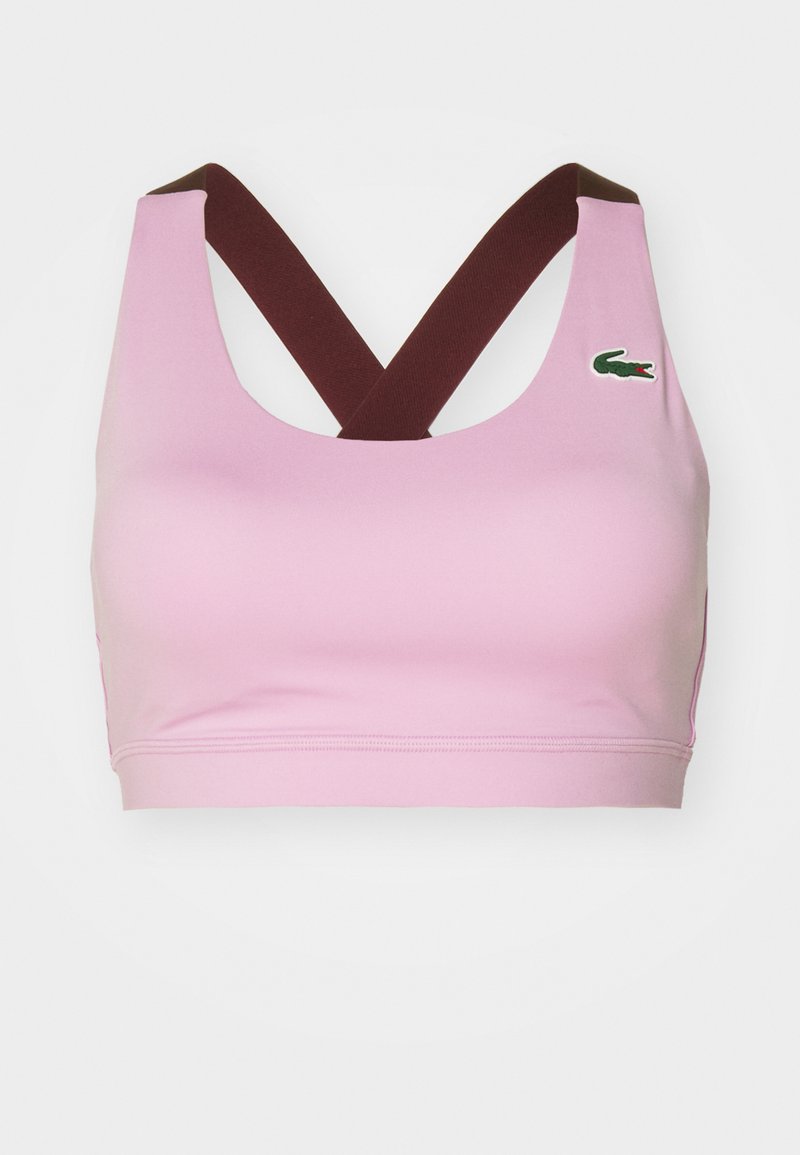 Sujetador deportivo rosa con amplias correas marrones entrelazadas y un pequeño logotipo de cocodrilo verde en la parte superior izquierda.