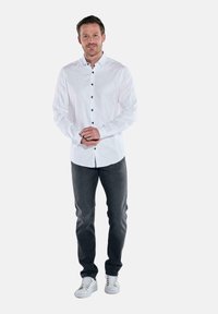 Mann steht mit verschränkten Händen, trägt ein weißes Hemd, dunkle Jeans und weiße Sneaker vor einem schlichten Hintergrund.