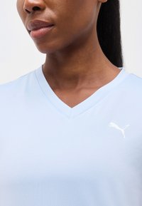 Κοντινό πλάνο ενός ατόμου που φορά ανοιχτομπλε μπλούζα με V-λαιμόκοψη και λευκό λογότυπο Puma στο στήθος, εμφανίζοντας τον λαιμό και το κάτω μέρος του προσώπου.