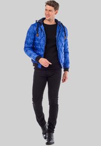 Blue quilted puffer jacket with a hood, black knit sweater, and slim black jeans. Features ribbed cuffs and detachable hood cords.
