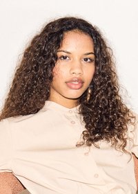 Jonge vrouw met lang krullend haar, dragend een beige hemd met knopen, kijkt recht vooruit met een neutrale uitdrukking tegen een effen achtergrond.