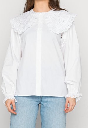 Femme portant un chemisier blanc avec un grand col et des poignets en dentelle à œillets, associé à un jean en denim bleu clair.