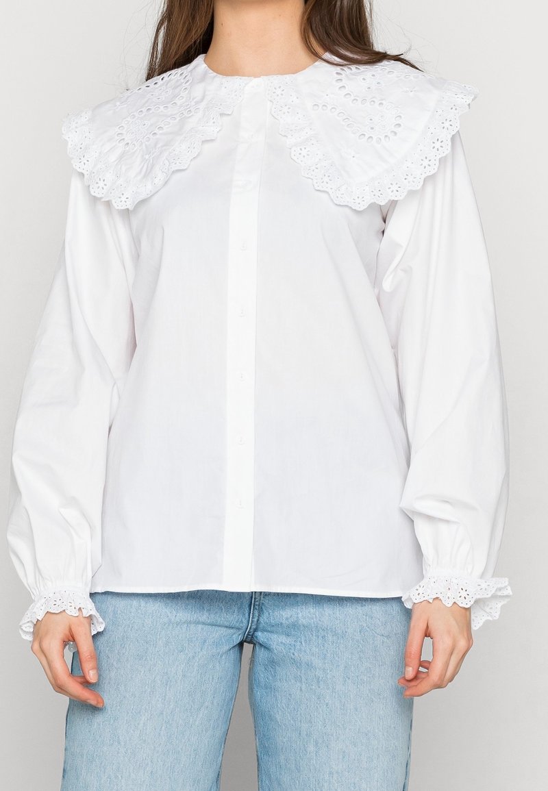 Femme portant un chemisier blanc avec un grand col et des poignets en dentelle à œillets, associé à un jean en denim bleu clair.