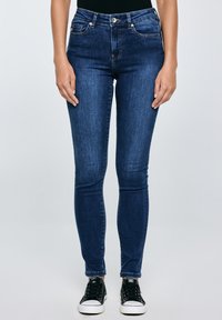 Femeie purtând blugi skinny albastru închis și adidași negri cu talpă joasă, stând pe un fundal simplu.