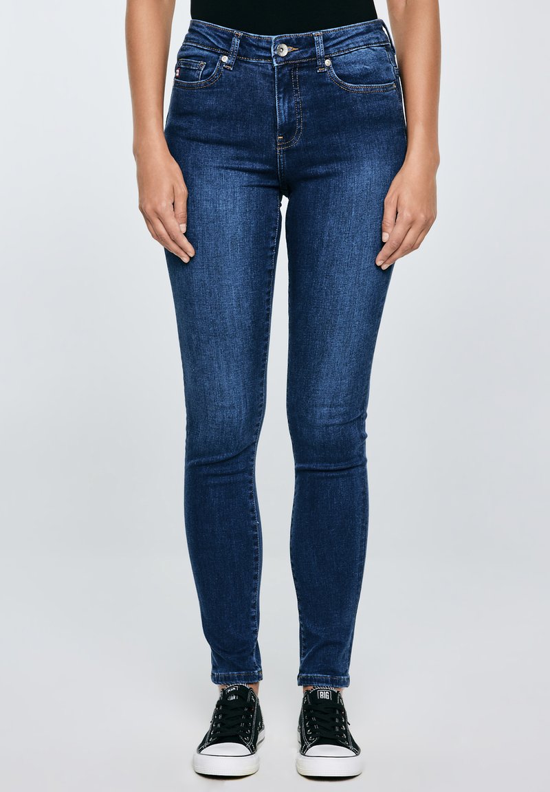 Femeie purtând blugi skinny albastru închis și adidași negri cu talpă joasă, stând pe un fundal simplu.