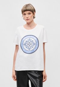 Λευκό βαμβακερό T-shirt με στρογγυλό μπλε γραφικό σχέδιο που απεικονίζει ένα χιόνι και στυλιζαρισμένο κείμενο. Στρογγυλή λαιμόκοψη, κοντά μανίκια, άνετη εφαρμογή.