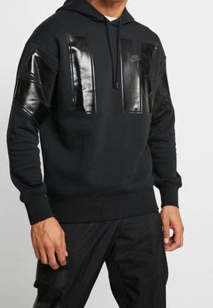 Mężczyzna w czarnej bluzie z kapturem Nike z błyszczącymi prostokątnymi naszywkami na klatce piersiowej i rękawach, w połączeniu z czarnymi spodniami z kieszeniami naszywanymi.