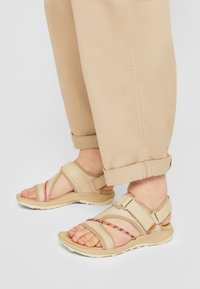 Beige sandaler har kryssremmer og en fargerik flettet detalj, med justerbare lukkinger og en polstret såle for komfort.
