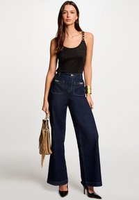 Top à bretelles noir avec un effet métallique, associé à un jean large taille haute bleu foncé avec poches et accents dorés. Escarpins noirs et sac à main beige.