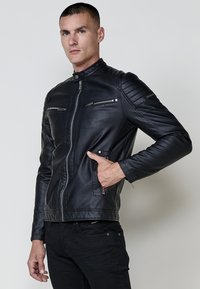Homme portant une veste en cuir zippée noire avec des détails matelassés aux épaules, une main dans la poche, se tenant contre un fond léger uni.