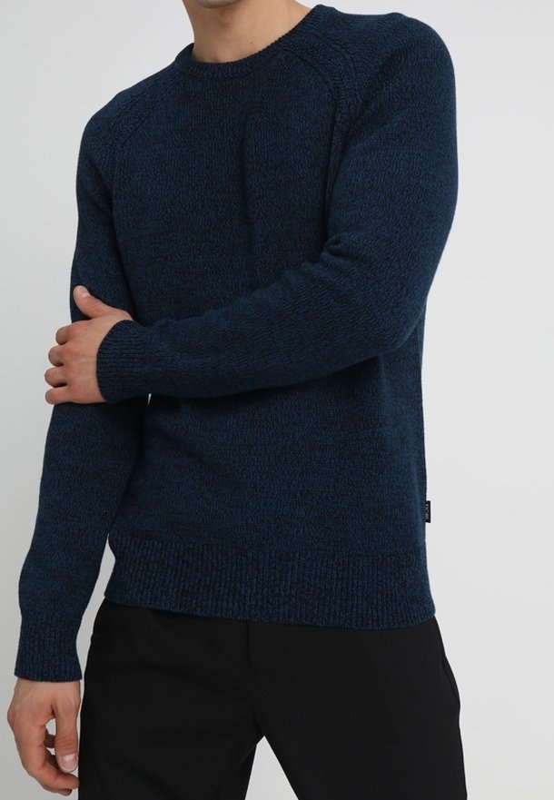 Man som bär en mörkblå strukturerad stickad tröja och svarta byxor, håller sin vänstra arm med sin högra hand mot en enfärgad bakgrund.