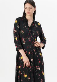 Svart blomstret kjole laget av lett stoff, med knappestolpe foran, lange ermer og flerfargede blomster mønstre gjennom hele.