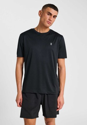 Młody mężczyzna stojący, ubrany w jednolity czarny T-shirt z krótkim rękawem z małym białym logo oraz pasujące czarne szorty na jednolitym tle.