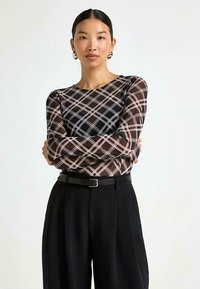 Långärmad, figursydd topp med rutigt mönster i bruna och svarta nyanser, gjord av ett genomskinligt material. Matchas med högmidjade svarta byxor och ett bälte.