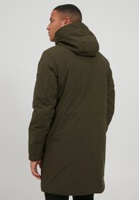 Chaqueta acolchada verde oliva con capucha, que presenta mangas largas y un cierre frontal con cremallera. Tejido suave con una textura ligeramente brillante y líneas limpias.