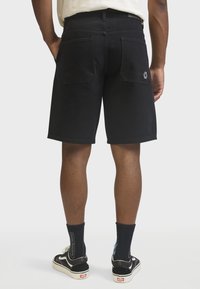 Homme portant un short noir, des chaussettes noires et des baskets noir et blanc, debout les mains dans les poches, vu de dos sur un fond uni.