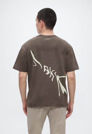 Άνδρας με την πλάτη, φοράει καφέ oversized T-shirt με μεγάλο ανοιχτό αφηρημένο σχέδιο κειμένου στην πλάτη και μπεζ παντελόνι.