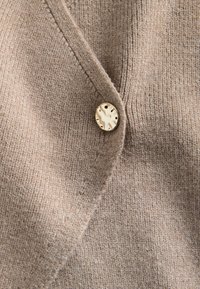 Beige geribbelde gebreide stof met een enkele ronde beige en zwarte gemarmerde knoop vastgemaakt door een knoopsgat.