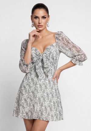 Sötét hajú nő fehér és szürke pöttyös ruhában, fodros ujjakkal és gyöngy fülbevalóval, az állát tartva pózol.