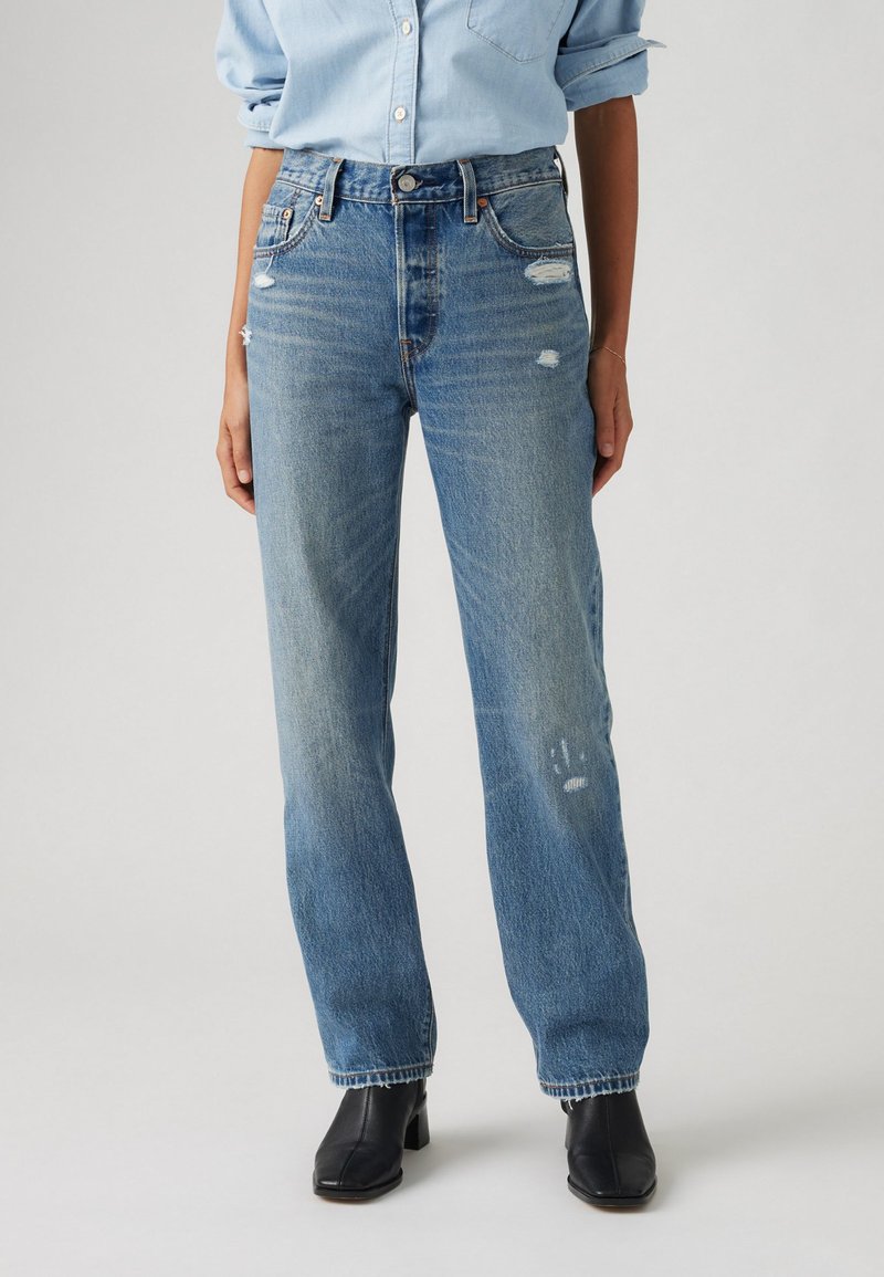 Jean en denim bleu clair avec une coupe droite, détails déchirés et légères marques d'usure, associés à des bottines noires.