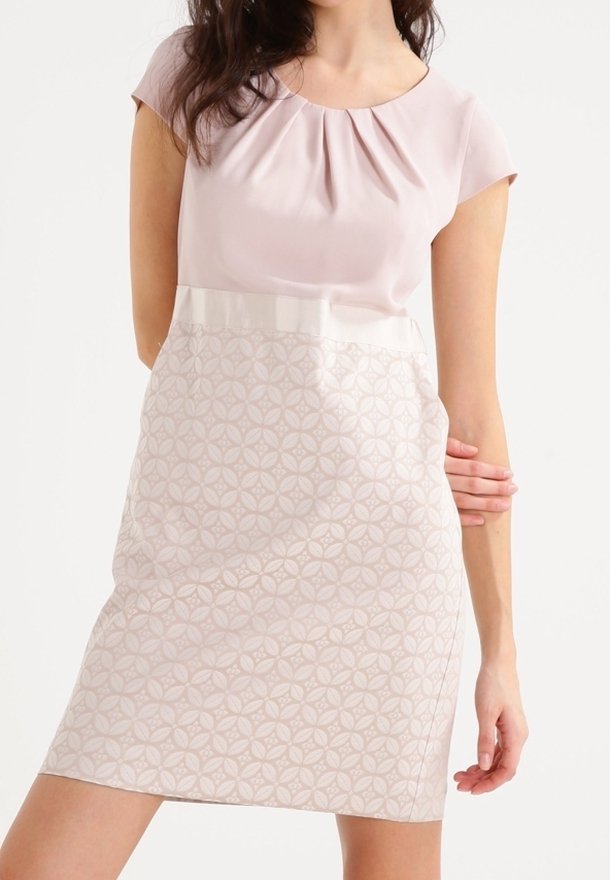 Femme portant une robe à manches courtes avec un haut rose uni et une jupe rose clair à motifs, posant avec un bras plié derrière le dos.
