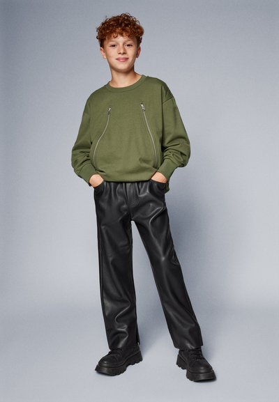 Sudadera verde con bolsillos con cremallera, combinada con pantalones de imitación de cuero negros y zapatillas negras de suela gruesa. El fondo es gris.