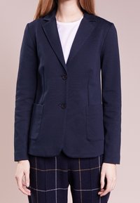Mujer vistiendo un blazer azul marino con dos botones, bolsillos de parche, camisa blanca y pantalones de cuadros oscuros con líneas claras.