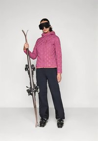 Mulher vestindo uma jaqueta de esqui rosa, calças pretas e botas de esqui, segurando esquis na posição vertical, usando grandes óculos de esqui pretos contra um fundo simples.