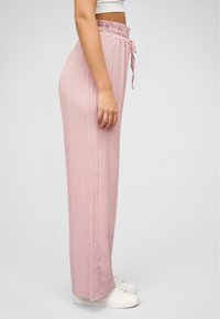Pantalons larges rose clair avec un design texturé à nervures. Comprennent une taille à cordon de serrage et une bande élastique. Portés avec des baskets blanches.