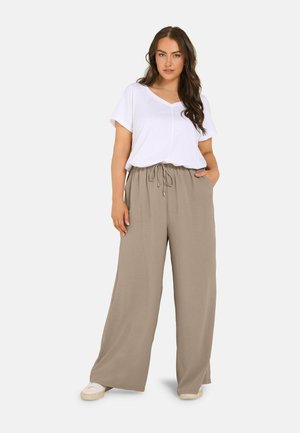 Jonge vrouw met lang haar, draagend een wit T-shirt met korte mouwen en losse taupekleurige wijdlopende broek met witte sneakers, staand met één hand in haar zak.