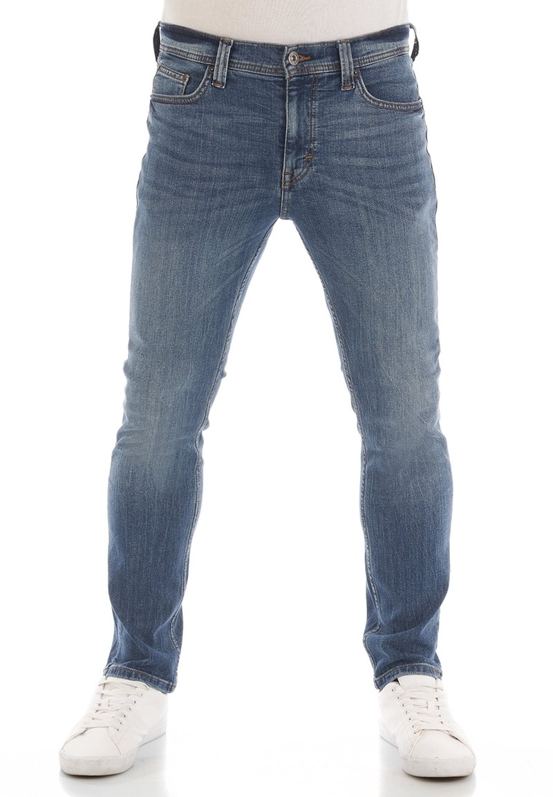 Homme portant un jean slim bleu et des baskets blanches, debout, jambes légèrement écartées sur un fond blanc.