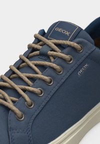Gros plan sur une basket bleue Geox avec des lacets beiges, des œillets métalliques, une étiquette de languette de marque et une semelle en caoutchouc blanche.