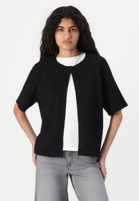 Black short-sleeve cardigan with a round neckline, single button closure, and soft texture, layered over a white shirt, paired with gray jeans.
