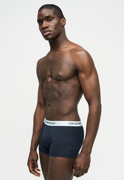 Muskularny mężczyzna stojący bokiem, ubrany w granatowe bokserki Emporio Armani z białym paskiem, na jednolitym jasnym tle.