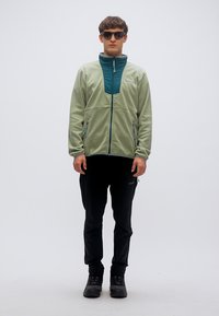 Φωτεινό πράσινο φλις μπουφάν με σκούρο τιρκουάζ γιακά, μπροστινό φερμουάρ και δύο πλαϊνές τσέπες, συνδυασμένο με μαύρο παντελόνι και σκουρόχρωμα παπούτσια.