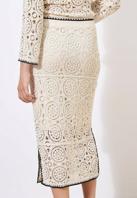 Jupe midi en dentelle crème avec des motifs circulaires, dotée d'une bordure noire le long de l' ourlet et de fentes latérales pour plus de détails et de texture.
