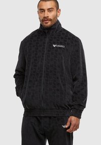 Homme portant une veste zippée noire texturée et un pantalon assorti, avec le logo "Colucci" sur la poitrine et le pantalon, se tenant devant un fond uni.