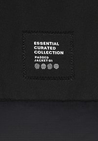 Schwarze gepolsterte Jacke mit einem aufgenähten Stoffetikett, das "Essential Curated Collection" und "Padded Jacket 01" zeigt, und darunter grafischen Mustern.