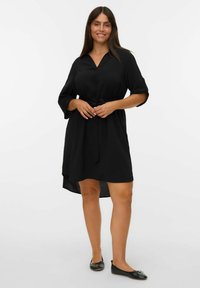 Zwarte jurk van lichtgewicht stof met een V-hals, driekwartmouwen en een gestrikt taille; asymmetrische zoom, gecombineerd met zwarte platte schoenen.