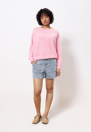 Light pink, textured knit sweater, loose fit. Blue denim shorts, front pockets. Brown flat shoes with ties. Model stands with hands in pockets.