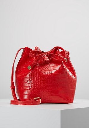 Rød spandtaske i kunstlæder med krokodillemønster, justerbar rem og snørelukning, vist på en hvid piedestal mod en ensfarvet baggrund.
