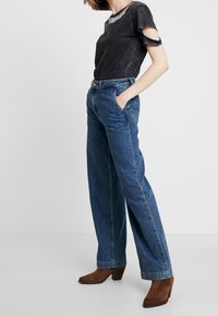 Camiseta de mezclilla negra con mangas cortadas, combinada con jeans azules de corte ancho y alta cintura, y botas de tacón marrones. Diseño sencillo, apariencia casual.