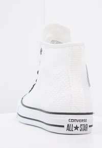 Bílé vysoké tenisky Converse All Star s texturovanou látkou, černým lemováním, kovovými očky a logem na patě na bílém pozadí.