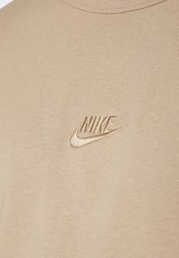 Béžové bavlněné tričko s jemně vyšitým logem Nike ve stejné barvě v horní části levého hrudníku. Hladká, dobře padnoucí textura.