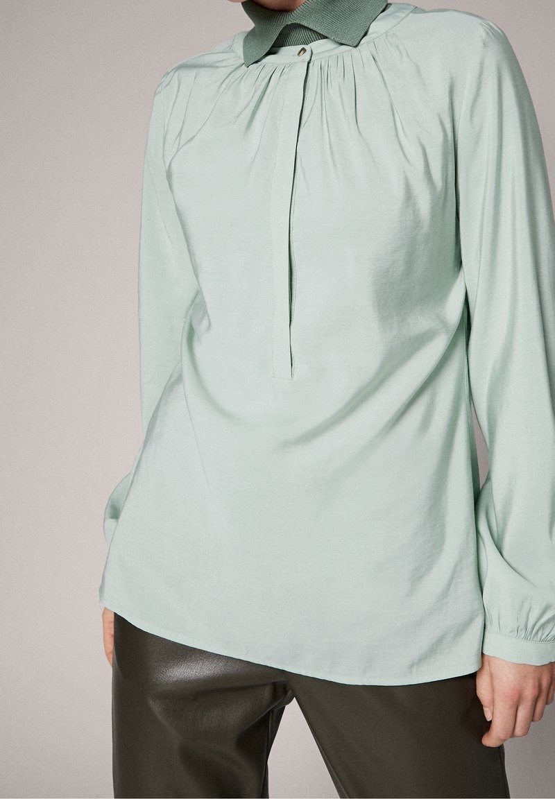 Personne portant un chemisier vert clair à manches longues avec des plis près du col et un pantalon vert olive, debout devant un fond neutre.
