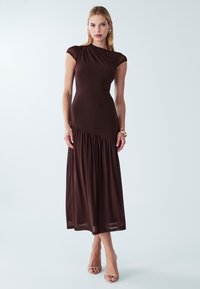 Žena stoji u smeđoj uskoj haljini s detaljem skupljenog dijela suknje, nosi zlatne naušnice u obliku obruča, narukvicu i bež otvorene štikle na bijeloj pozadini.