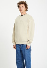 Beige sweatshirt made of cotton-blend fabric, with a crew neckline and embossed "VOLCOM" logo on the chest. Paired with blue denim pants.